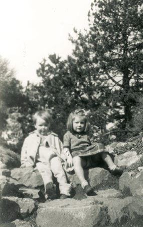 A young Evelyn Doyle sitting on the rock with his brother while wearing a checkered dress and black shoes