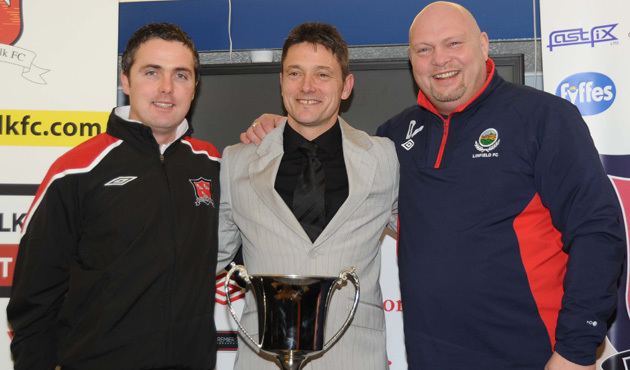 Dessie Gorman and the two men beside him are smiling with a trophy in front of them. Dessie is wearing a black long sleeve under a black necktie and gray coat. The man on the left is wearing a black and red jacket while the bald man with beard on the right is wearing a blue and red jacket