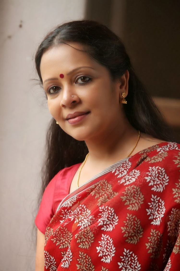 Devi Ajith smiling and wearing a red saree, dangling earrings and necklace with a red mark on the forehead