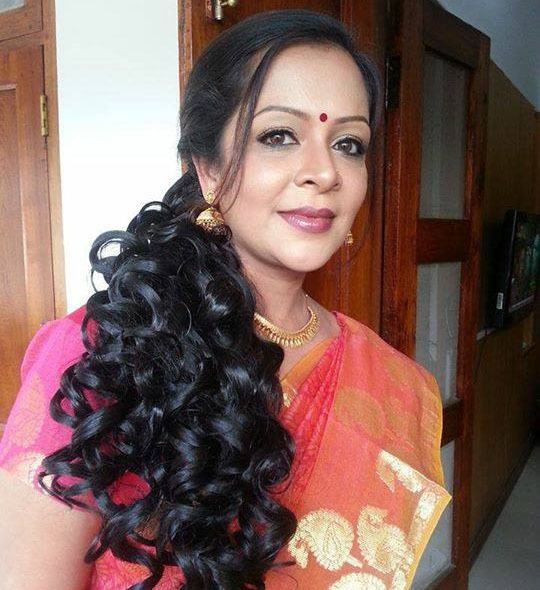 Devi Ajith smiling with curly hair and a red mark on her forehead while wearing a pink saree, dangling earrings, and necklace
