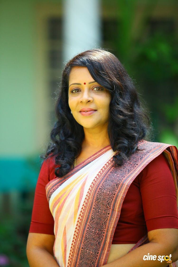 Devi Ajith smiling  while wearing a red saree with a red mark on the forehead