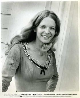 Diana Ewing smiling, with curly blonde hair, and wearing a long-sleeve dress with a black ribbon.