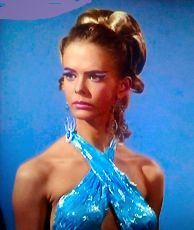 Diana Ewing as Droxine with a serious face, blonde hair, wearing chandelier earrings, and a sexy blue top in a scene from Star Trek (1966–1969 film).