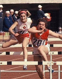 Dianne Gerace wearing a red headband, a sleeveless shirt, and red shorts while doing high jump together with her opponent.