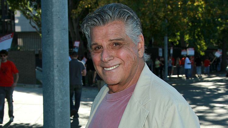 Dick Gautier smiling with people in the background having gray hair and wearing an off-white coat over a pink shirt.