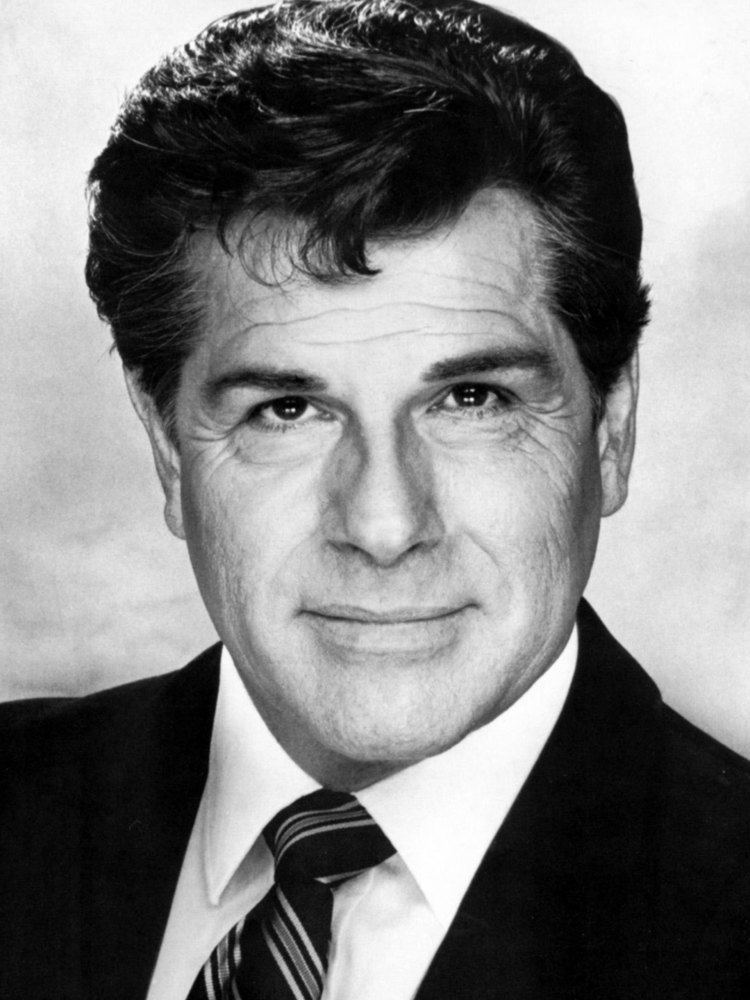 Dick Gautier with a tight-lipped smile, wearing a black coat over white long sleeves and a black and white striped necktie.