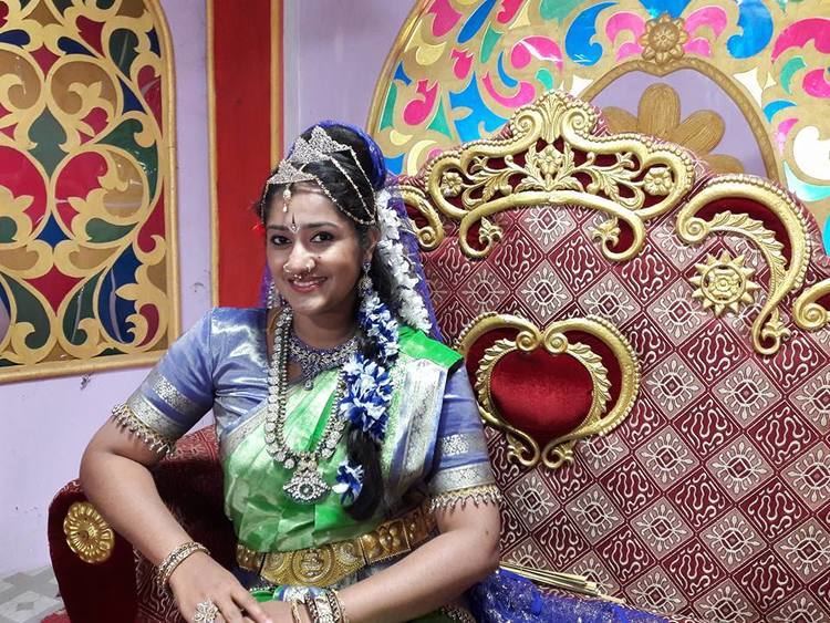 Dini Daniel sitting down on a red and gold bench and smiling while wearing green and blue Sri Lankan dress and more jewelry.