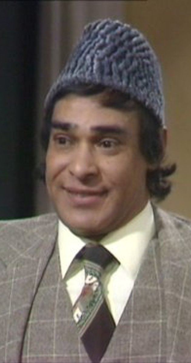 Dino Shafeek smiling while wearing a gray checkered coat, vest, yellow long sleeves, and necktie in a scene from the 1977 tv series, Mind Your Language