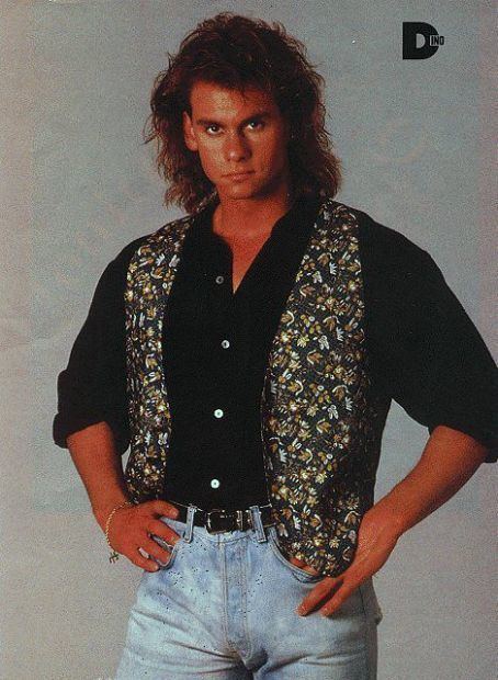 Dino hands on his hips while wearing black long sleeves, a floral vest, a black belt, and denim pants