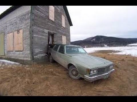 An Oldsmobile Custom Cruiser standing in front of a small wooden house with snow in the background.