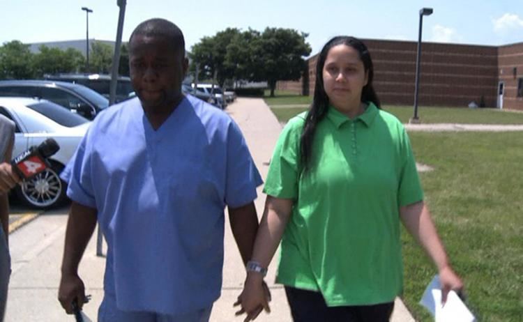 Charlie Bothuell IV (left) is seriously walking with his wife, has black hair, on his right are cars parked, and a microphone, right hand holding his keys, left hand holding the hand of her wife, he is wearing a blue top and pants. Monique Dilliard-Bothuell (right) is serious, has black hair, her left hand holding a paper, her right hand holding the hand of her husband, and wearing a wristwatch on her right hand and a green shirt and black pants.