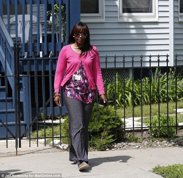 A woman wearing a Michigan Department of Human Services badge is walking away, has black hair, her left hand holding a phone, her right hand wearing a red bracelet, a colored top with a diamond design, a pink blazer, gray pants, and gray slippers.