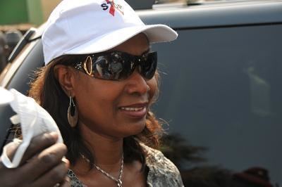 Djene Kaba Condé smiling while looking afar and wearing a white cap, black sunglasses, blouse, earrings, and necklace