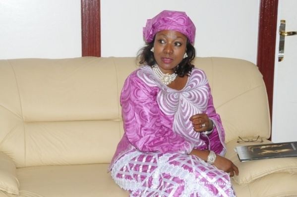 Djene Kaba Condé sitting on the couch while looking at something and wearing a violet bandana, white and violet long dress, and some pieces of jewelry