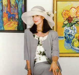 Dolores Erickson smiling while wearing a hat, a gray blazer, floral blouse, and gray skirt