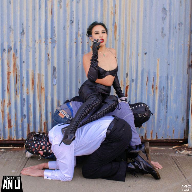 Two men kneeling on the floor while a woman sitting on their back, smoking and wearing a black brassiere, skirt, and high boots which signifies Dominatrix