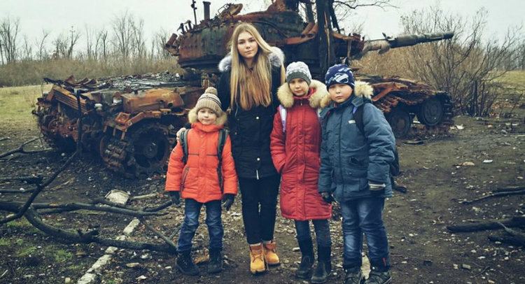 Maryana Naumova with a tight-lipped smile together with three little boys with serious faces in front of a decayed warrior armored vehicle. Maryana with long blonde hair, wearing a black coat, black pants, and brown shoes while the three boys are wearing bonnets and coats.