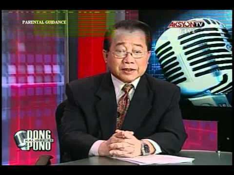 Dong Puno while reporting on Aksyon TV, with a serious face while his hands are on the table, wearing eyeglasses, a watch, a black coat over white long sleeves, and a brown tie.