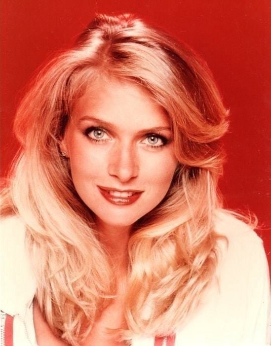 Donna Dixon smiling with wavy blonde hair while wearing a white blouse in a red background