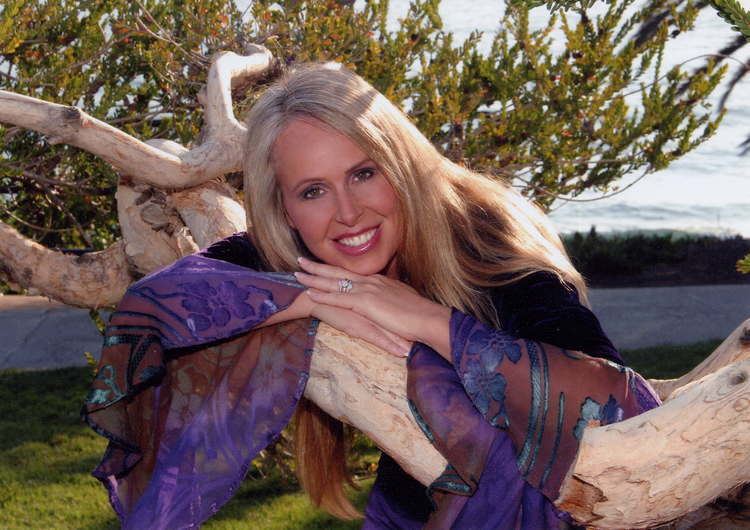 Doreen Virtue smiling while leaning on the tree and wearing a black blouse and blue and brown scarf