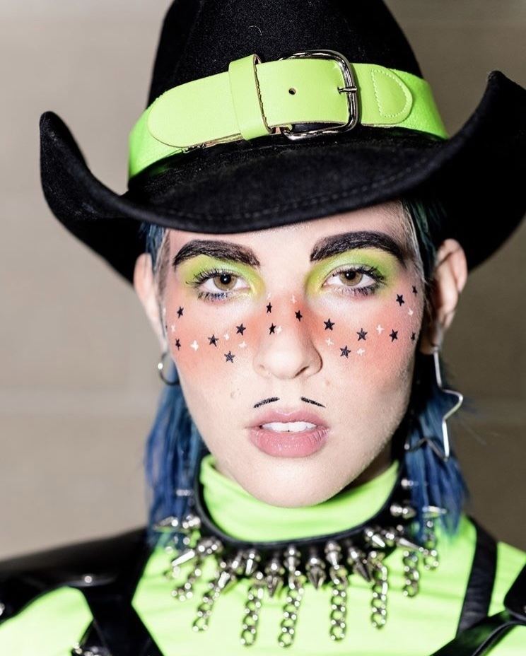 Dorian Electra with stars on her cheeks, neon green eyeshadow, and blue hair while wearing, black hat, chocker, and black and neon green blouse