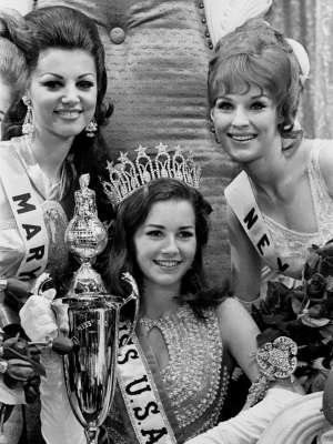 Dorothy Anstett smiling with her co-beauty queens during Miss USA 1968 coronation night. Dorothy holding a trophy and a scepter, wearing a crown and a sash.