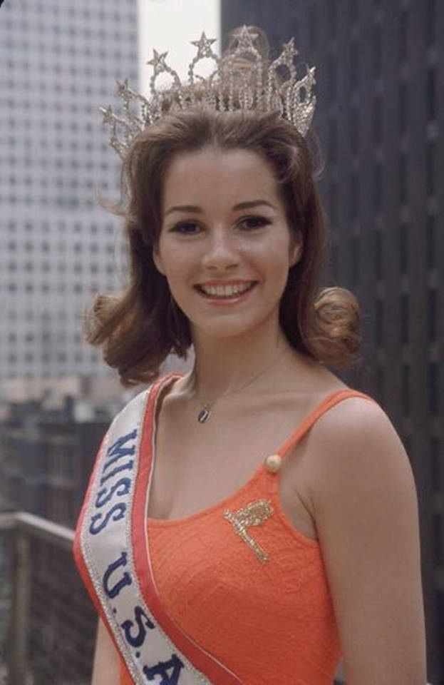 Miss USA 1968 Dorothy Anstett smiling, with wavy blonde hair, wearing a crown, a sash, a necklace, and an orange spaghetti shirt.