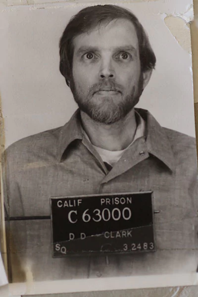 A younger Doug Clark Douglas with thick facial hair posing for his mugshot wearing a prison inmate attire.