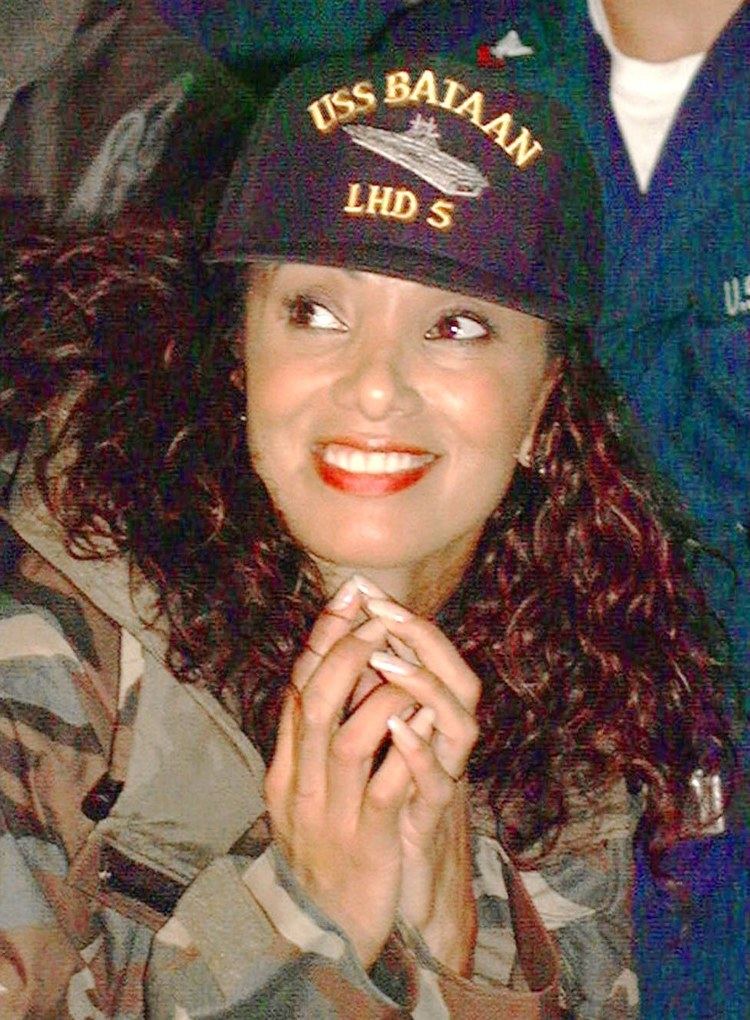 Downtown Julie Brown with a smiling face, with curly hair, wearing a cap and an army fatigue jacket.
