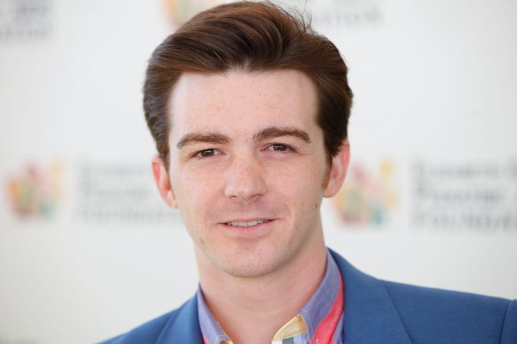 Drake Bell smiling while wearing a colored shirt under a blue coat