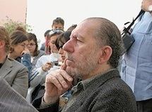 Driss El Khouri in a serious face looking at something in a side view with his right hand touching his chin having a beard and mustache with people in the background and wearing a gray tweed wool coat.