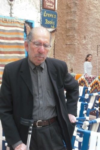 Driss El Khouri is looking serious with a beard and mustache while holding a cane and taking out something from his pocket and there are chairs, printed cloth, and a girl in the background. Driss is wearing eyeglasses and wearing a pair of black pants with a brown belt, and a gray collared shirt under a black coat.