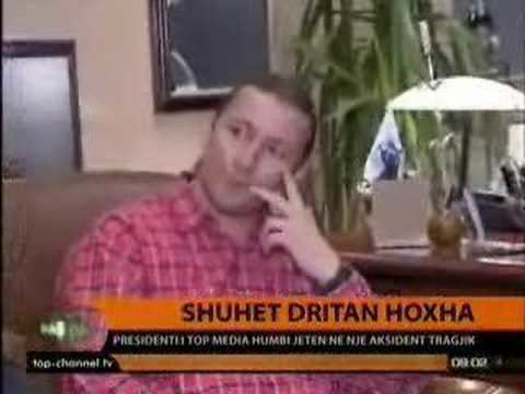 A clip from a TV telecast on the tragic accident and lost of Dritan Hoxha, an Albanian businessman and the founder of Top Media, Albania's largest media company. In the image, Dritan is in an office setting in the background, with short brown hair, has two fingers on his left cheek, and a wristwatch on his left, wearing a red checkered long sleeve.