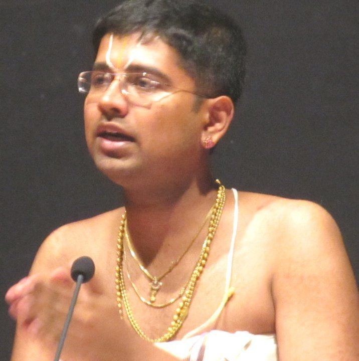 Dushyanth Sridhar talking into a microphone while wearing eyeglasses and necklaces