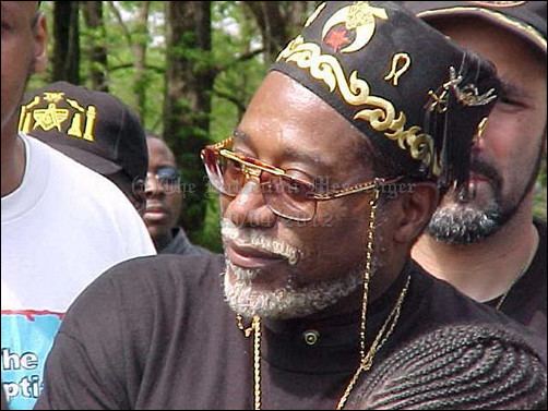 Dwight York with a tight-lipped smile, mustache, and beard while wearing a black and gold cap, black t-shirt, eyeglasses, and necklace