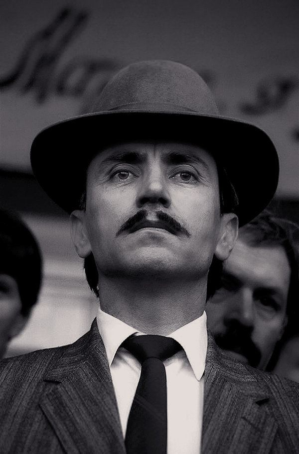 Dzhokhar Dudayev looking afar, with a serious face and mustache with a man on his background, while wearing a hat and a white long sleeve under a necktie and striped coat
