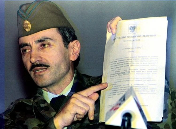 Dzhokhar Dudayev presented a document to the press on a microphone, with a serious face and mustache, while wearing a brownish-green side cap with a gold pin and a white long sleeve under a black sweater and camouflage jacket