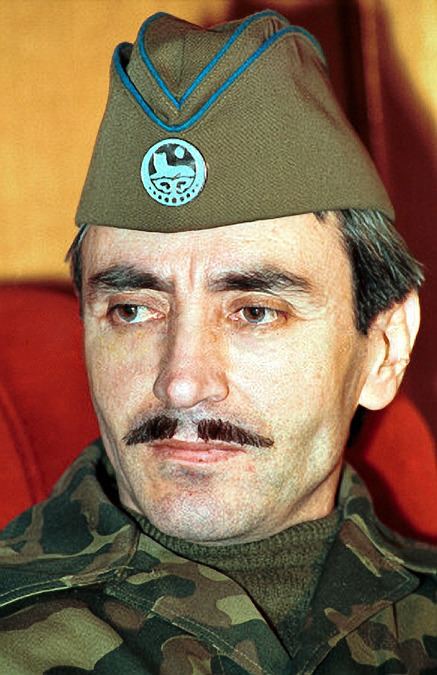 Dzhokhar Dudayev looking at something, with a serious face and mustache, while wearing a brownish-green side cap with a pin and a brownish-green shirt under a camouflage jacket