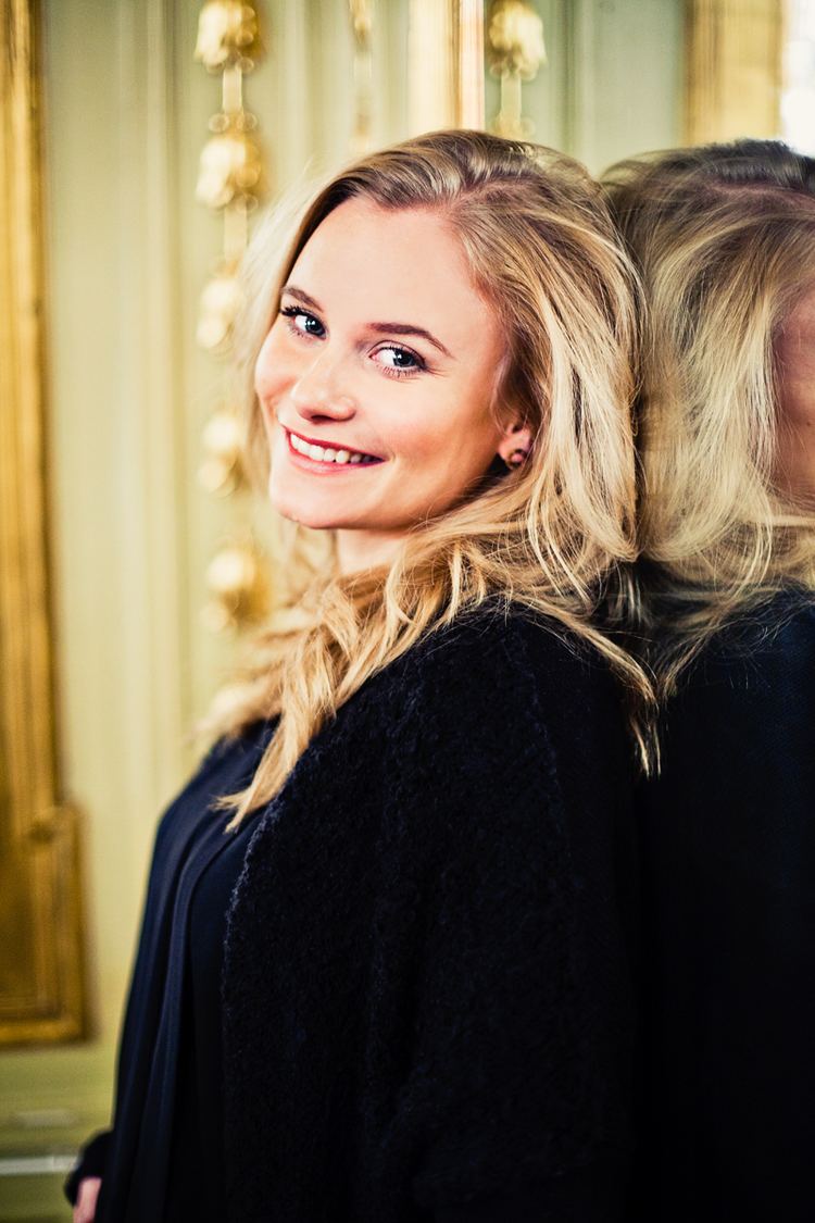 Ebba Hultkvist with a smiling face while leaning in the mirror, with blonde wavy hair, and wearing a black long sleeve top.