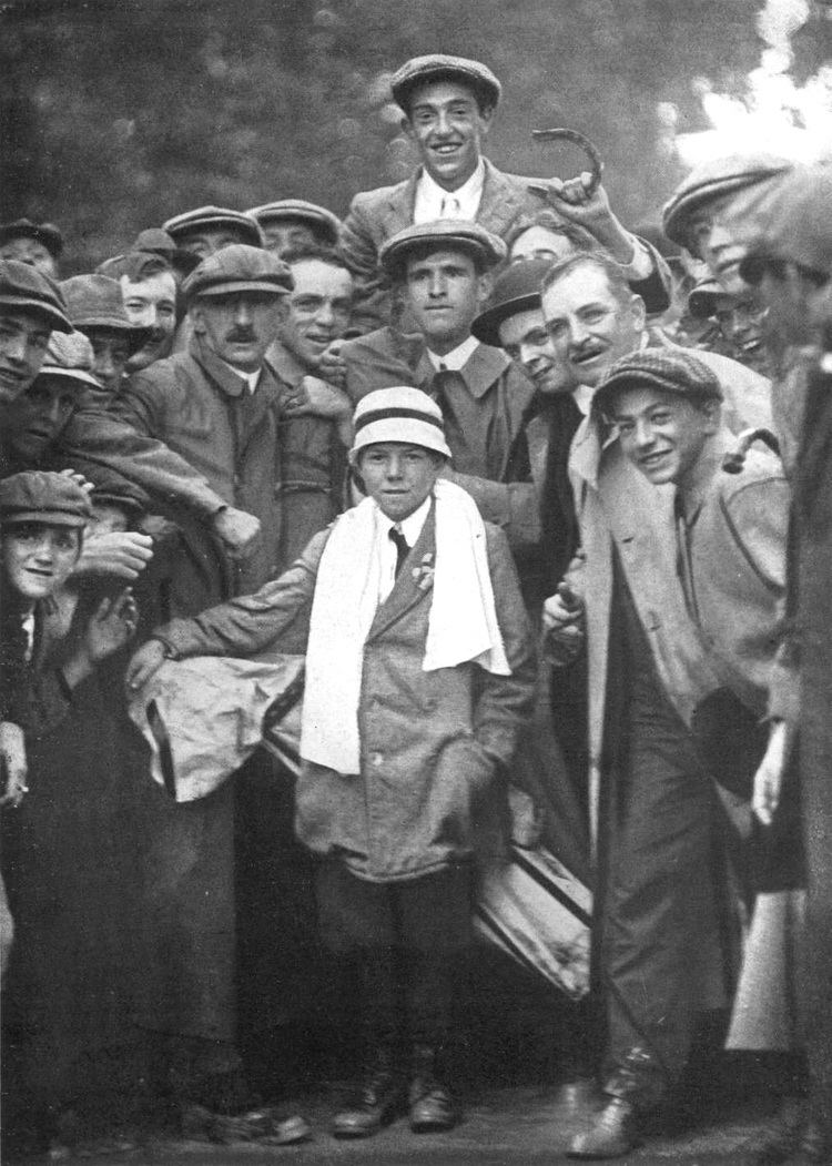 In an iconic photograph of Eddie Lowery and Francis Ouimet together along with a golfer and their audience, in the middle is Eddie Lowery smiling, standing with his left hand in his pocket while holding a golf club bag on his right shoulder, has black hair, wearing a white hat with black line, towel on around his neck, white long sleeves with black necktie and coat, at the top is Francis Ouimet, happy, has black hair wearing golf driving cap white shirt with necktie and a coat.