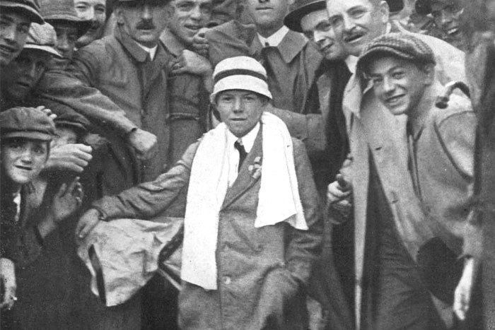 In an iconic photograph of Eddie Lowery and Francis Ouimet together along with a golfer and their audience, in the middle is Eddie Lowery smiling, standing with his left hand in his pocket while holding a golf club bag on his right shoulder, has black hair wearing a white hat with black line, towel on around his neck, white shirt with black necktie and coat.
