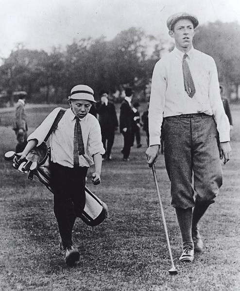 In an iconic photograph of Eddie Lowery and Francis Ouimet striding down the fairway together, along with four gentlemen at their back wearing a white bowtie, a necktie, black coat, and black and white pants, on the left is Eddie lowery serious looking down holding a white golf club bag on his right shoulder while walking, wearing a white cap, white polo with black necktie, black shoes, and a black pants, at the right Francis Ouimet is serious, looking straight, holding a golf club on his right hand, left hand down, wearing a white long sleeve polo with black necktie, black shoes, and black pants.