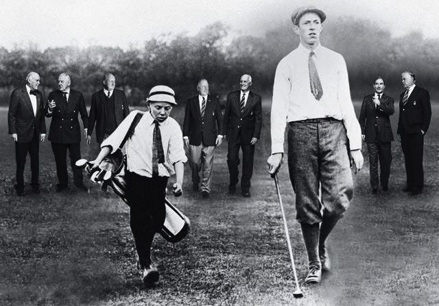 An iconic photograph of Eddie Lowery and Francis Ouimet striding down the fairway together, along with a seven gentleman at their back wearing white with bowtie, a necktie, black coat, black and white pants, on the left is Eddie lowery serious looking down holding a white golf club bag on his right shoulder while walking, wearing white cap, white polo with black necktie, black shoes and a black pants, at the right Francis Ouimet is serious, looking straight, holding a golf clubs on his right hand, left hand down, wearing a white long sleeve polo with black necktie, black shoes and black pants.