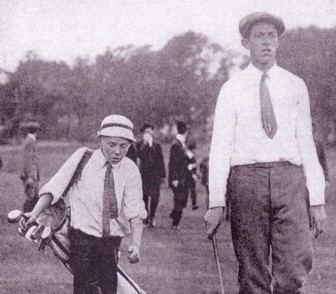 An iconic photograph of Eddie Lowery and Francis Ouimet striding down the fairway together, along with four gentlemen at their back wearing white with bowtie, a necktie, black coat, and black and white pants, on the left is Eddie lowery, serious, looking down holding a white golf club bag on his right shoulder while walking, wearing a white cap, white polo with a black necktie and a black pants, at the right Francis Ouimet is serious, looking straight, holding a golf club on his right hand, left hand down, wearing a white long sleeve polo with a black necktie and black pants.