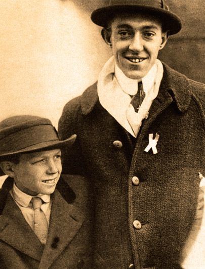 Eddie lowery is smiling, standing looking up to his left, has black hair, wearing a hat, white polo with tie, and black coat, at the right Francis Ouimet is smiling, standing beside Eddie, hands down behind Eddie’s shoulder, has black hair wearing a black hat, white long sleeves with black tie, and black coat.