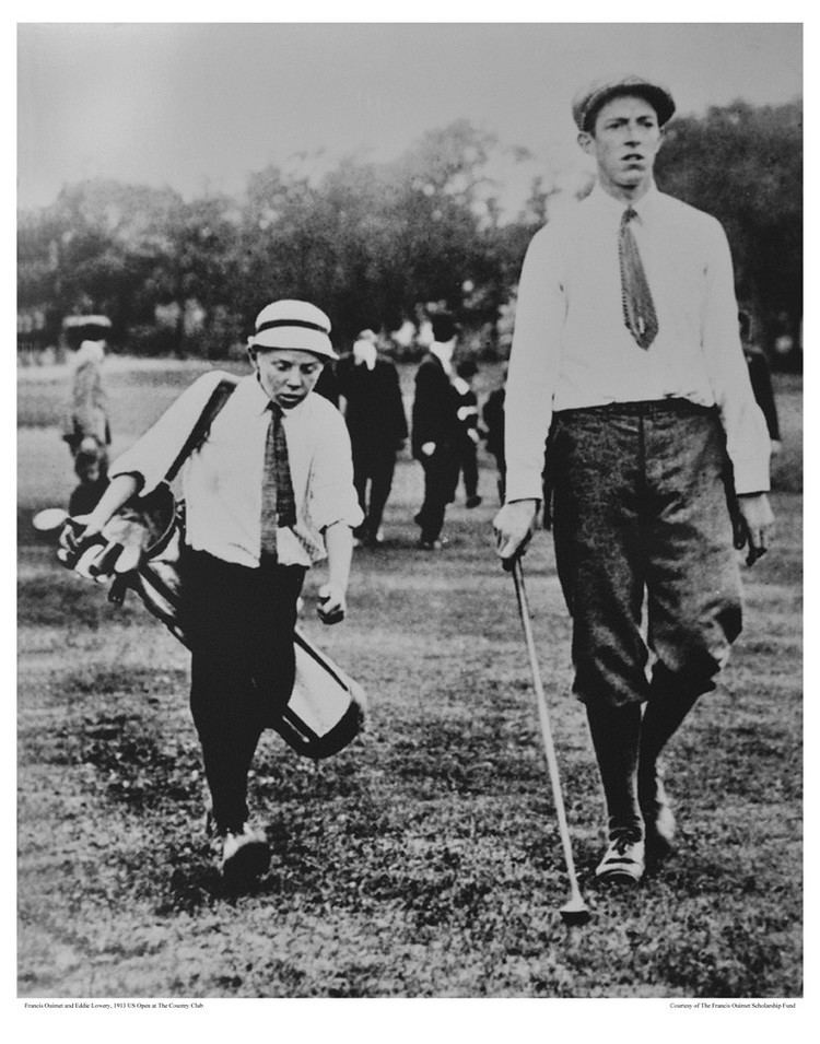 In an iconic photograph of Lowery and Ouimet striding down the fairway together, along with four gentlemen at their back, wearing a white bowtie, a necktie, black coat, and black and white pants, on the left is Eddie lowery serious looking down holding a white golf club bag on his right shoulder while walking, wearing a white cap, white polo with black necktie, black shoes, and a black pants, at the right Francis Ouimet is serious has, looking straight, holding a golf club on his right hand, left hand down, wearing a white long sleeve polo with black necktie, black shoes and black pants.