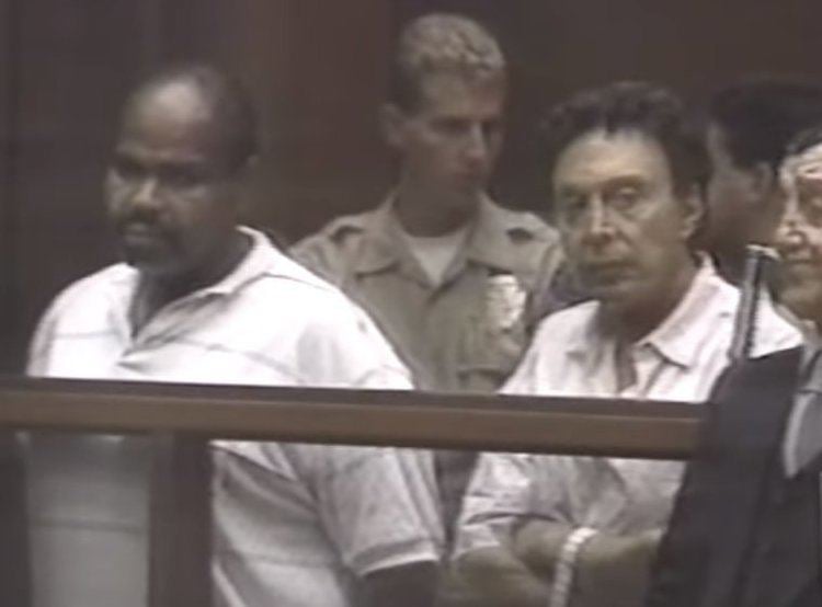 Gregory Diles on left is serious, bald, and has a mustache, he is wearing a white with a black lined shirt, Eddie Nash on right is serious, has black curly hair, both hands crossed, wearing a wristwatch on his left hand, and white long sleeves. A man in front of Eddie Nash is serious, has black hair, wearing dark gray long sleeves under a black suit. Behind Gregory (on left) is a policeman, serious, has curly hair, wearing a white shirt under a brown police uniform and a badge on his left chest, and on right is a man, who has black hair, talking to the policeman.