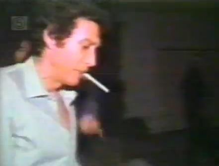 Eddie Nash is serious, has black curly hair, a cigarette in his mouth, he is wearing light blue long sleeves.