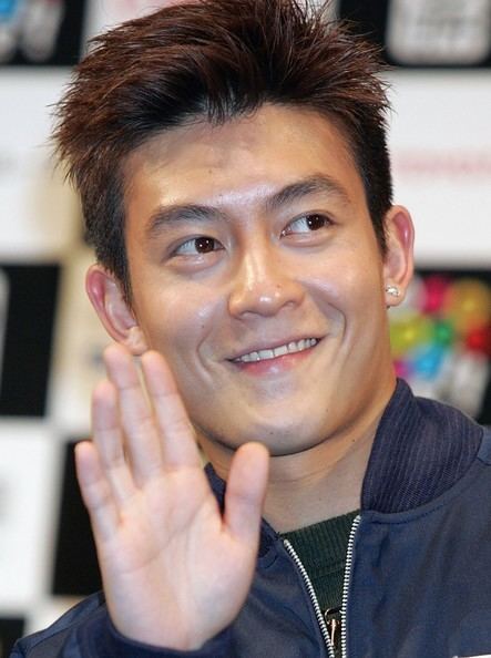 Edison Chen waving and wearing a green shirt under a blue jacket
