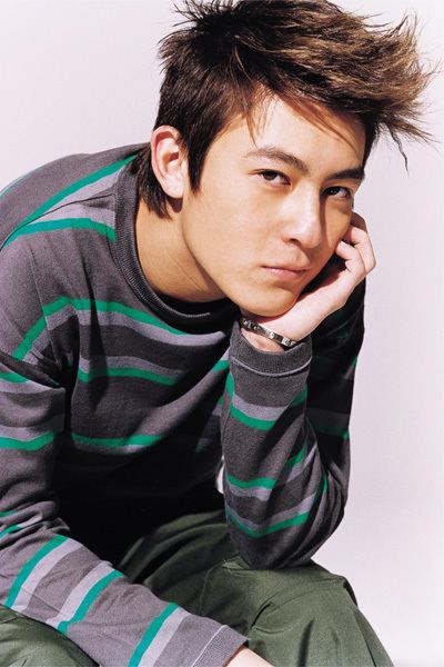 Edison Chen looking serious with left hand on his cheek and wearing a black, green, and purple jacket and gray pants